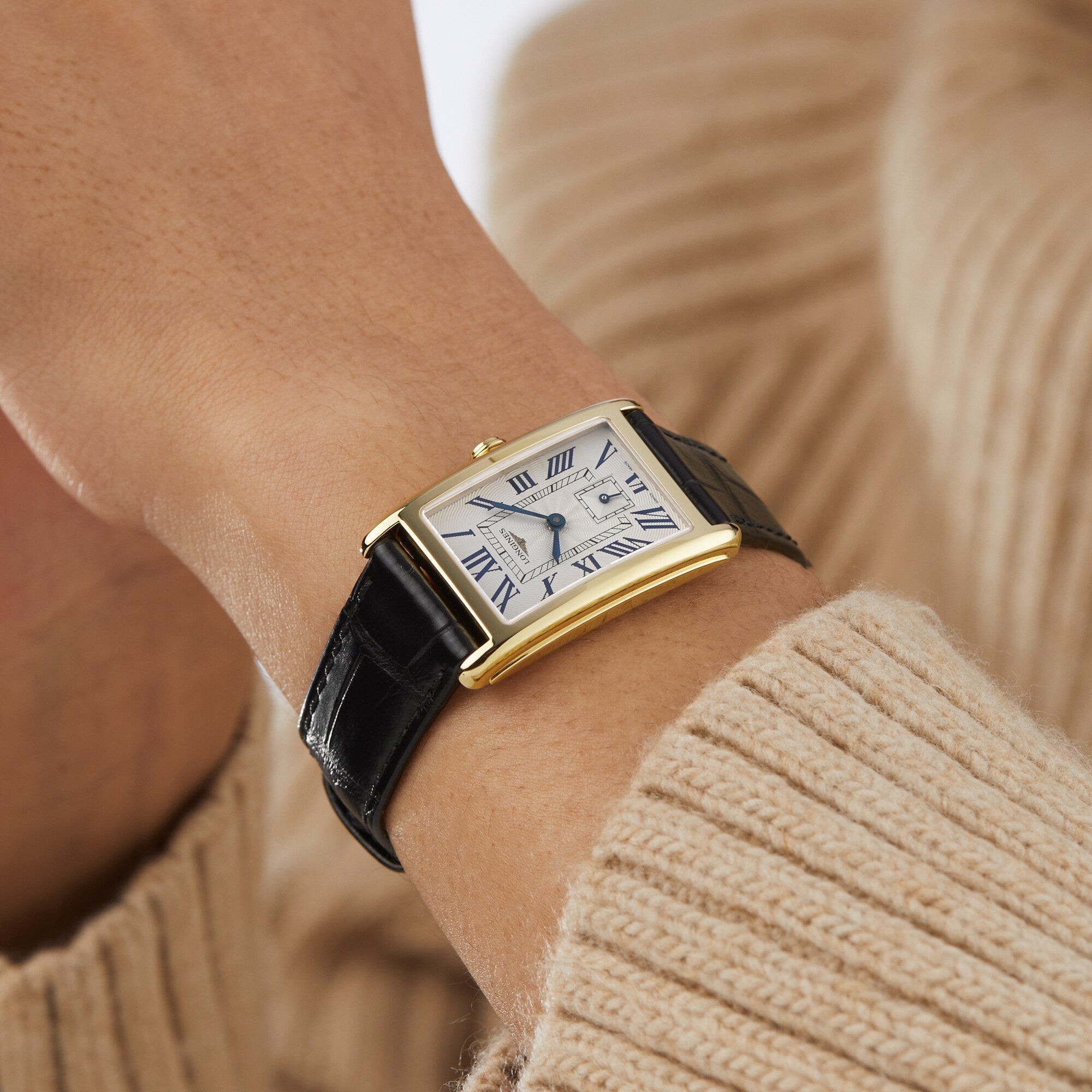 A Longines DolceVita watch with a rectangular gold case, white dial, black Roman numerals, and a black leather strap highlights classic style on the wrist of someone in a beige sweater.