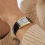 A Longines DolceVita watch with a rectangular gold case, white dial, black Roman numerals, and a black leather strap highlights classic style on the wrist of someone in a beige sweater.