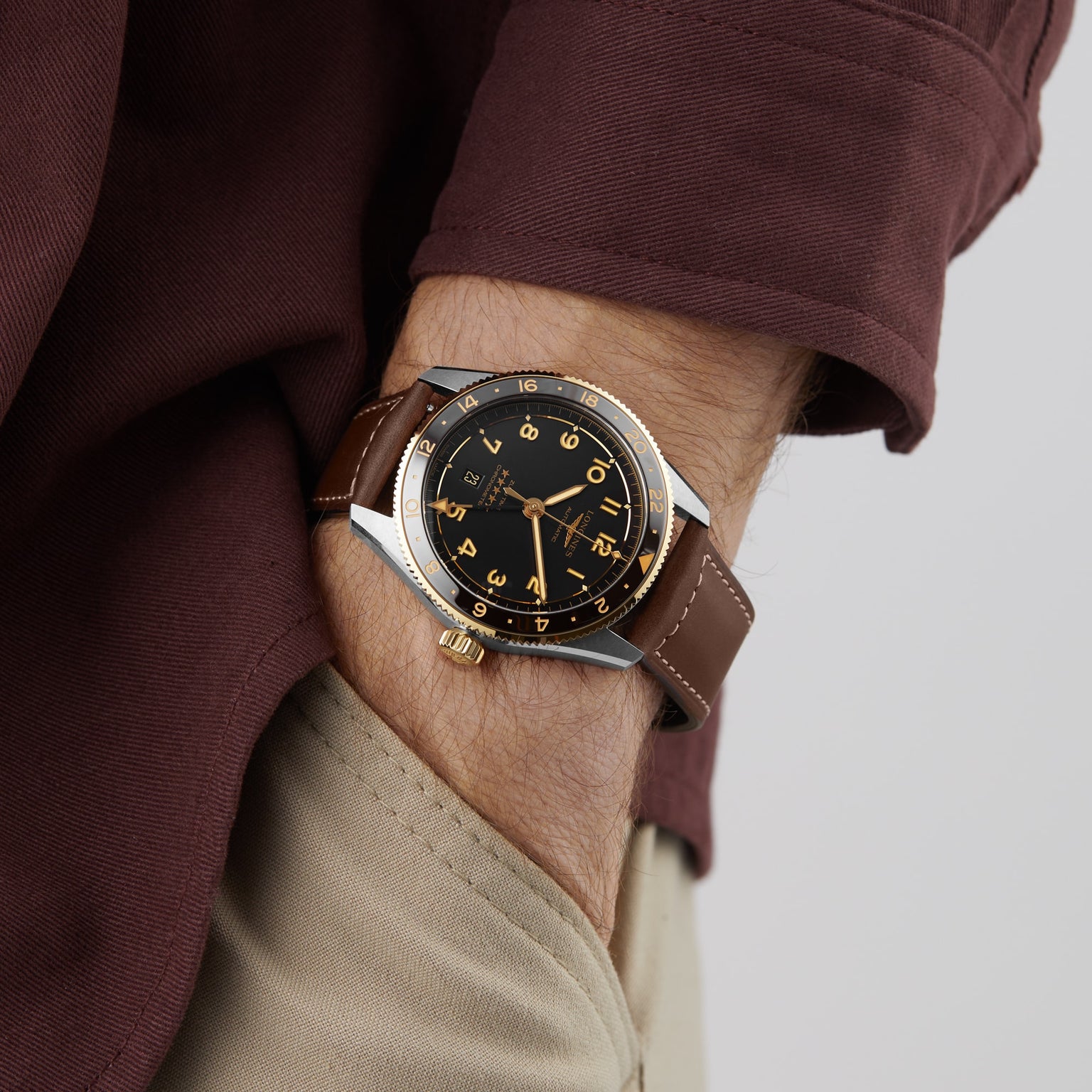 A person in a brown long-sleeve shirt and beige pants wears a Longines Spirit Zulu Time watch with a black and gold face on their left wrist, capturing the spirit of exploration.