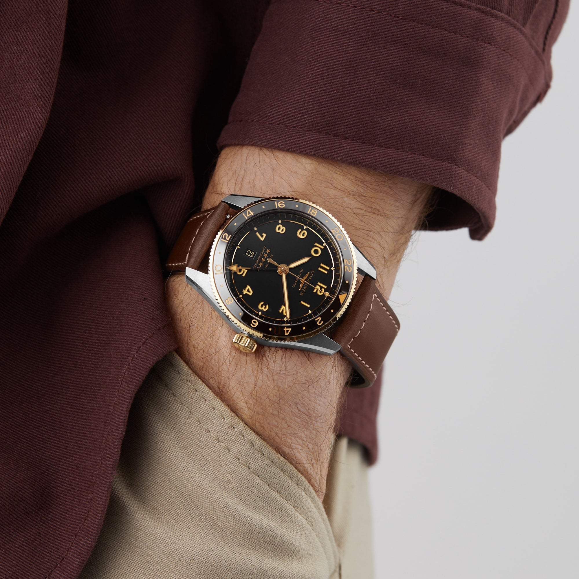 A person in a brown long-sleeve shirt and beige pants wears a Longines Spirit Zulu Time watch with a black and gold face on their left wrist, capturing the spirit of exploration.