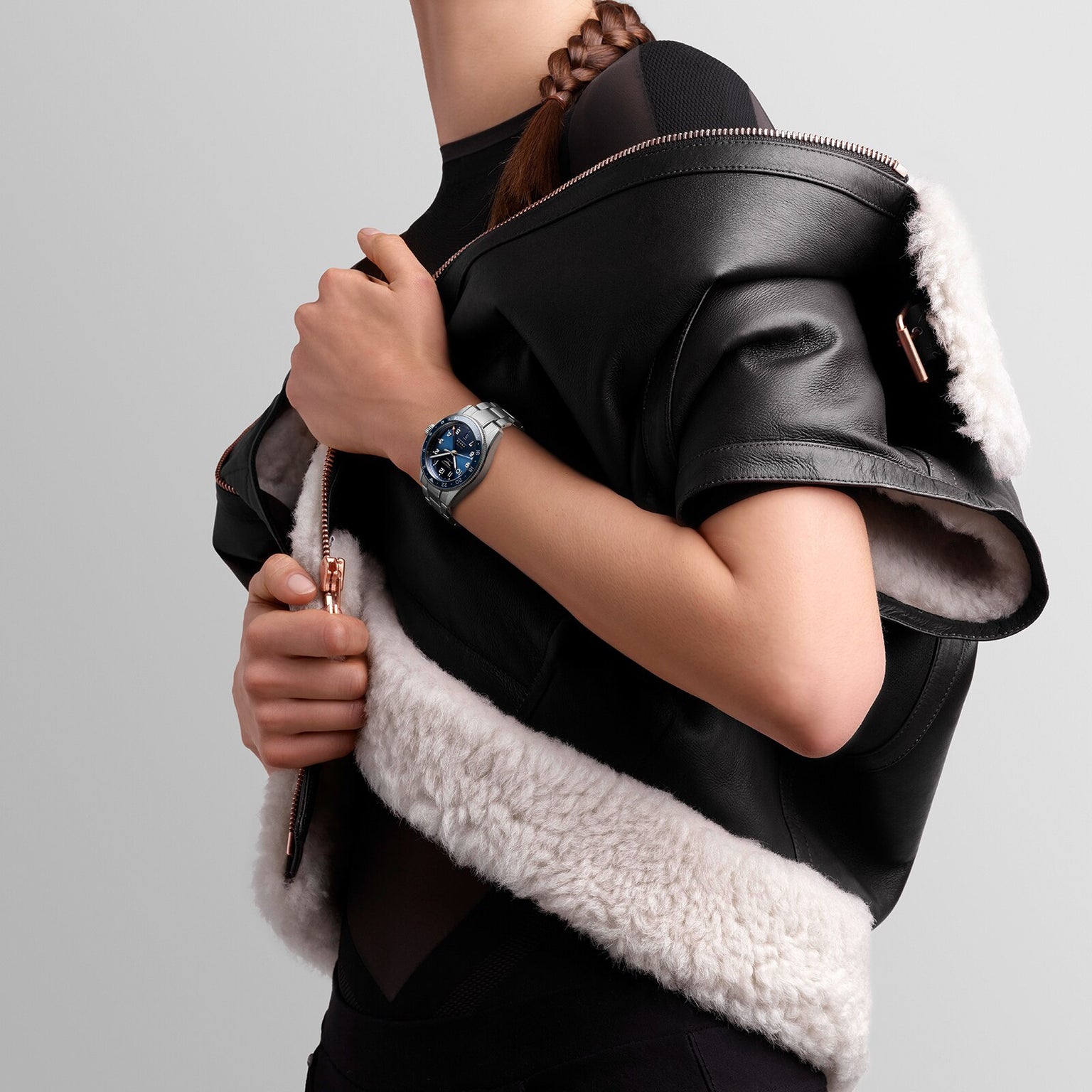 A person with a black and white shearling jacket draped over their shoulders reveals a Longines Spirit Zulu Time watch on their left wrist, featuring the distinctive design of the renowned Swiss brand.