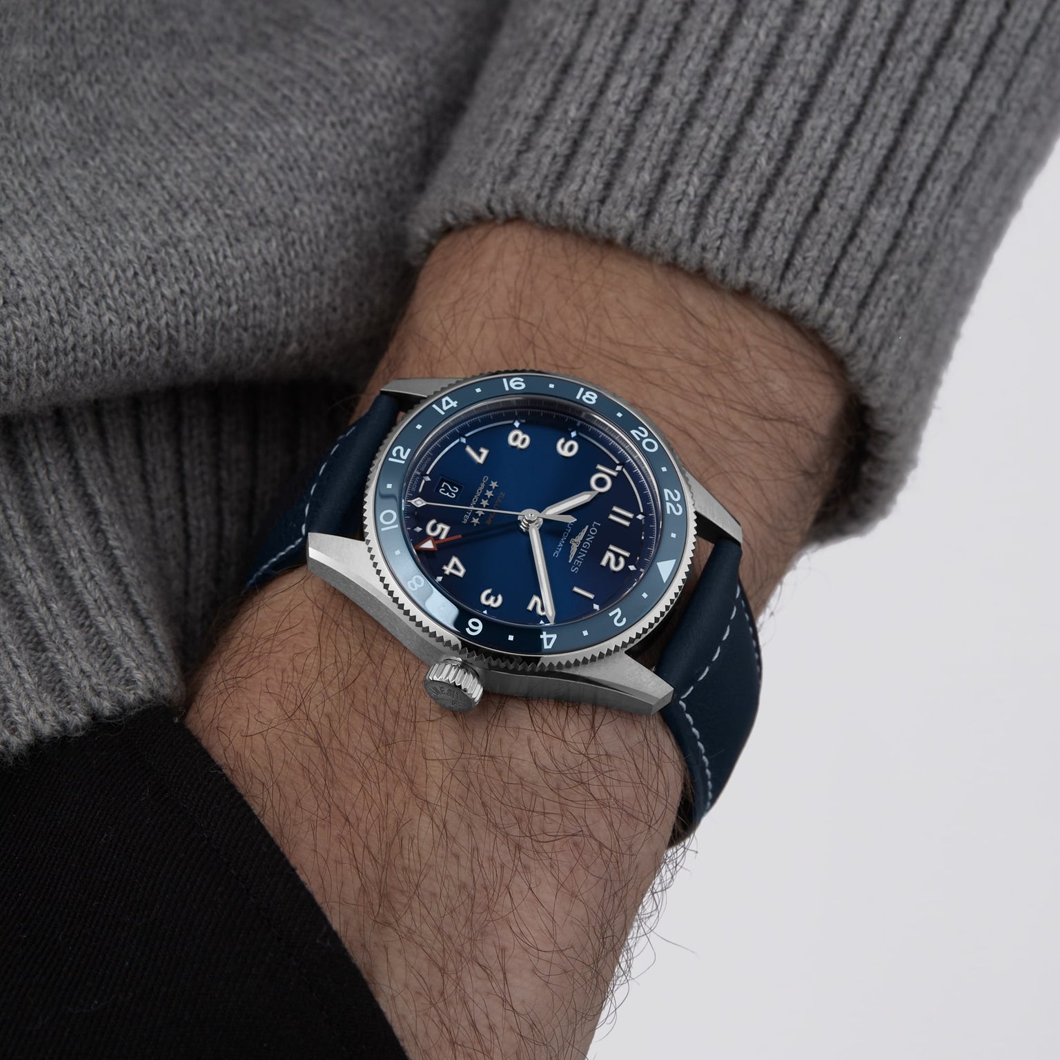A person in a gray sweater showcases the Longines Spirit Zulu Time watch, featuring a blue dial, blue leather strap, and stainless steel case on their left wrist.
