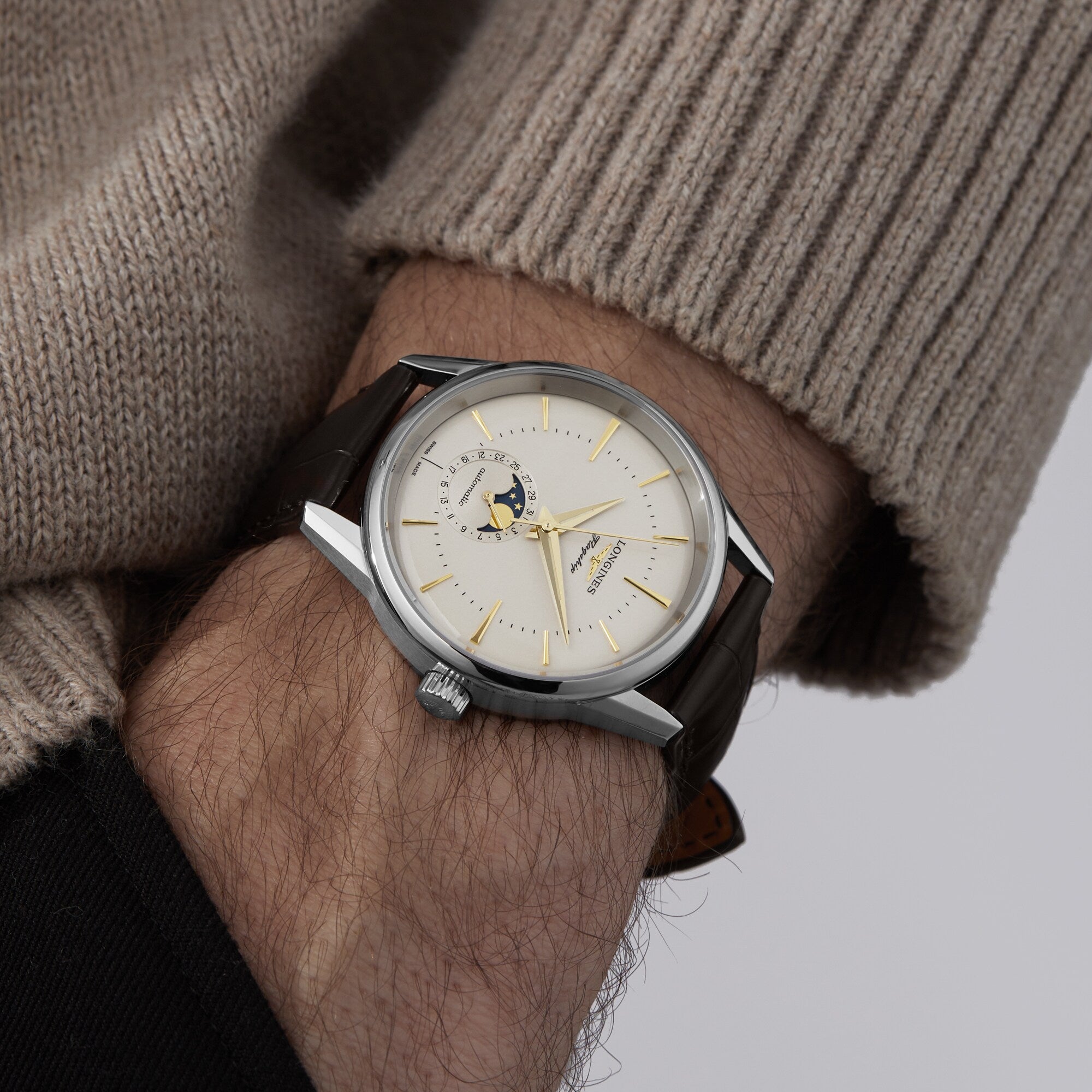 A person in a beige sweater is showcasing the Longines Flagship Heritage Moonphase wristwatch, featuring a cream dial, gold hour markers, a moon phase display, and a brown leather strap.