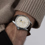 A person in a beige sweater is showcasing the Longines Flagship Heritage Moonphase wristwatch, featuring a cream dial, gold hour markers, a moon phase display, and a brown leather strap.