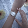 With manicured nails, someone wears a Longines Master Collection Moonphase silver watch featuring a white dial, moon phase indicator, and diamond indexes, displayed on marble with yellow flowers.
