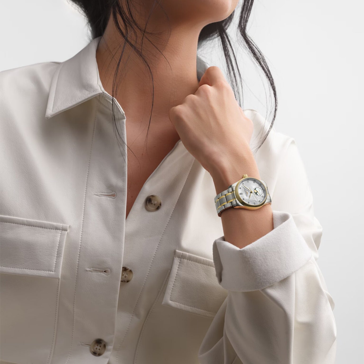 A woman in a cream button-up poses with her left hand raised, showcasing the Longines Master Collection Moonphase watch with a white dial, gold accents, and elegant diamond hour markers.