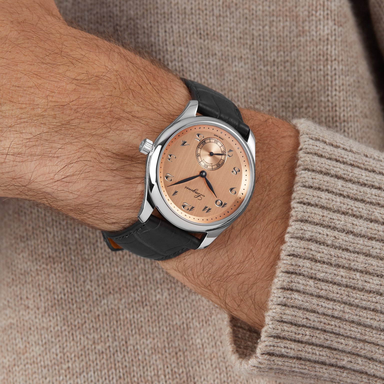 A person in a beige sweater showcases the Longines Master Collection wristwatch with a salmon pink dial, Roman numerals, and a black leather strap.