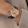 A person in a beige sweater showcases the Longines Master Collection wristwatch with a salmon pink dial, Roman numerals, and a black leather strap.