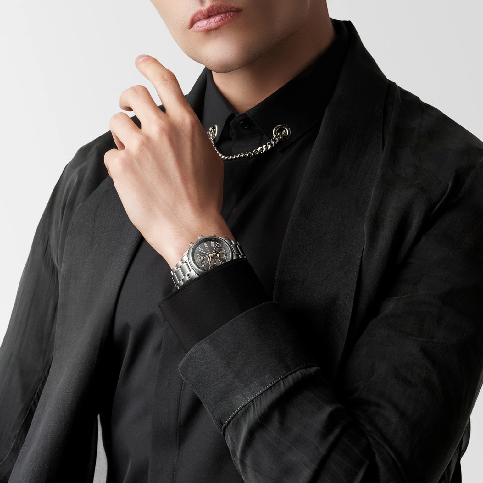 A person in a black suit and shirt, wearing the Longines Master Collection Chrono Moonphase watch and a chain necklace, poses with one hand near their face against a plain background.