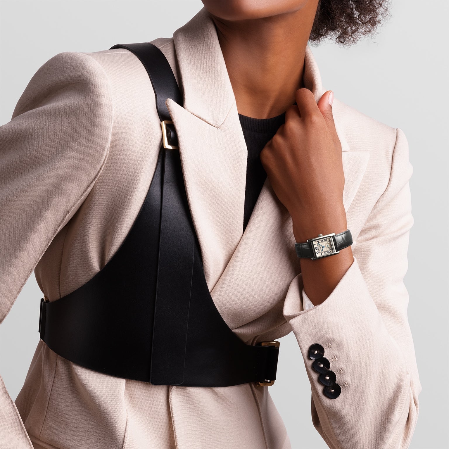 A person in a cream blazer with black shoulder straps wears a Longines Mini DolceVita with a rectangular stainless steel case on their left wrist, posing with one hand by the collar.