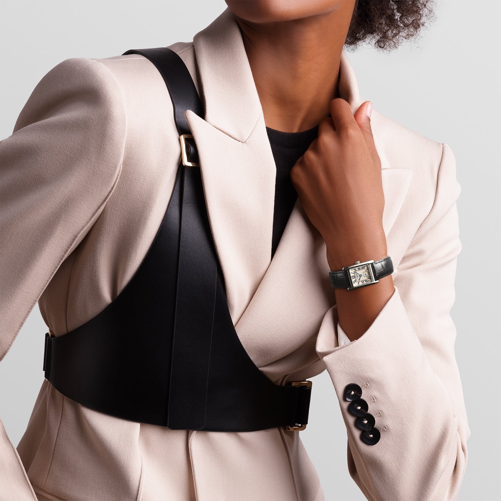 A person in a cream blazer with black shoulder straps wears a Longines Mini DolceVita with a rectangular stainless steel case on their left wrist, posing with one hand by the collar.