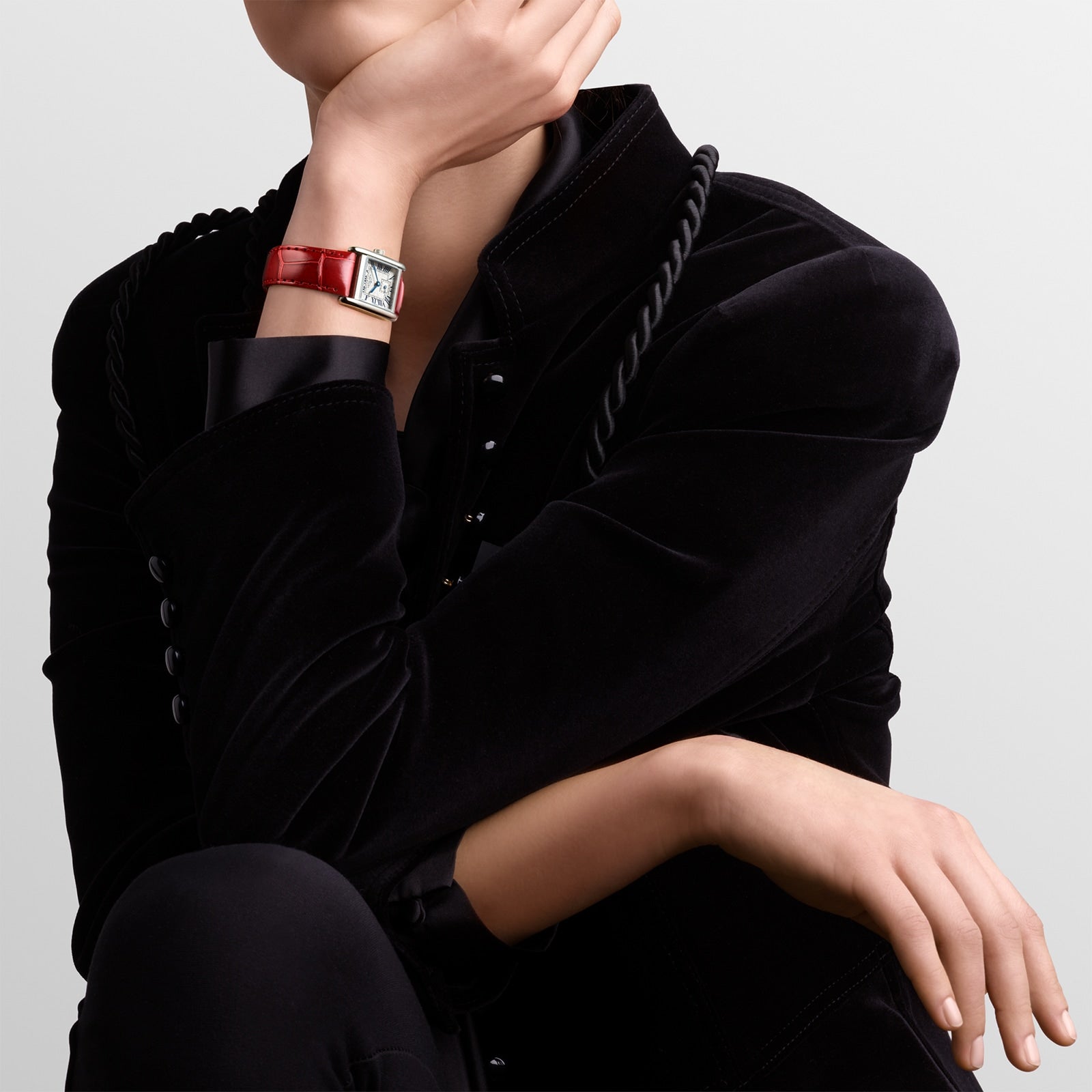 Person in a black velvet jacket wearing a Longines Mini DolceVita red leather-strapped wristwatch, resting their chin on their hand against a plain light background.