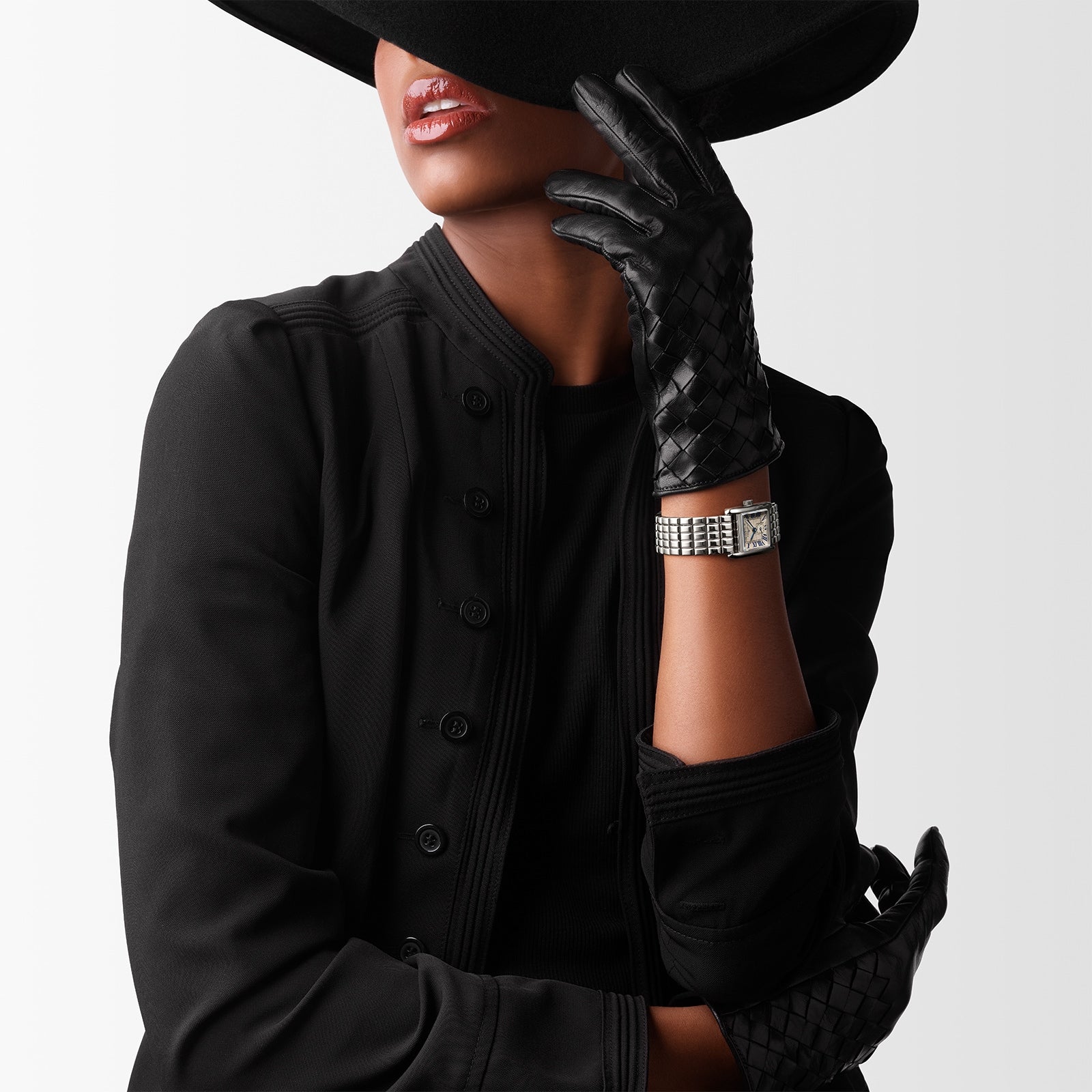 A woman in black attire and gloves poses with a wide-brim hat shading her eyes, showcasing the Longines Mini DolceVita—a luxury watch that exudes contemporary sophistication.