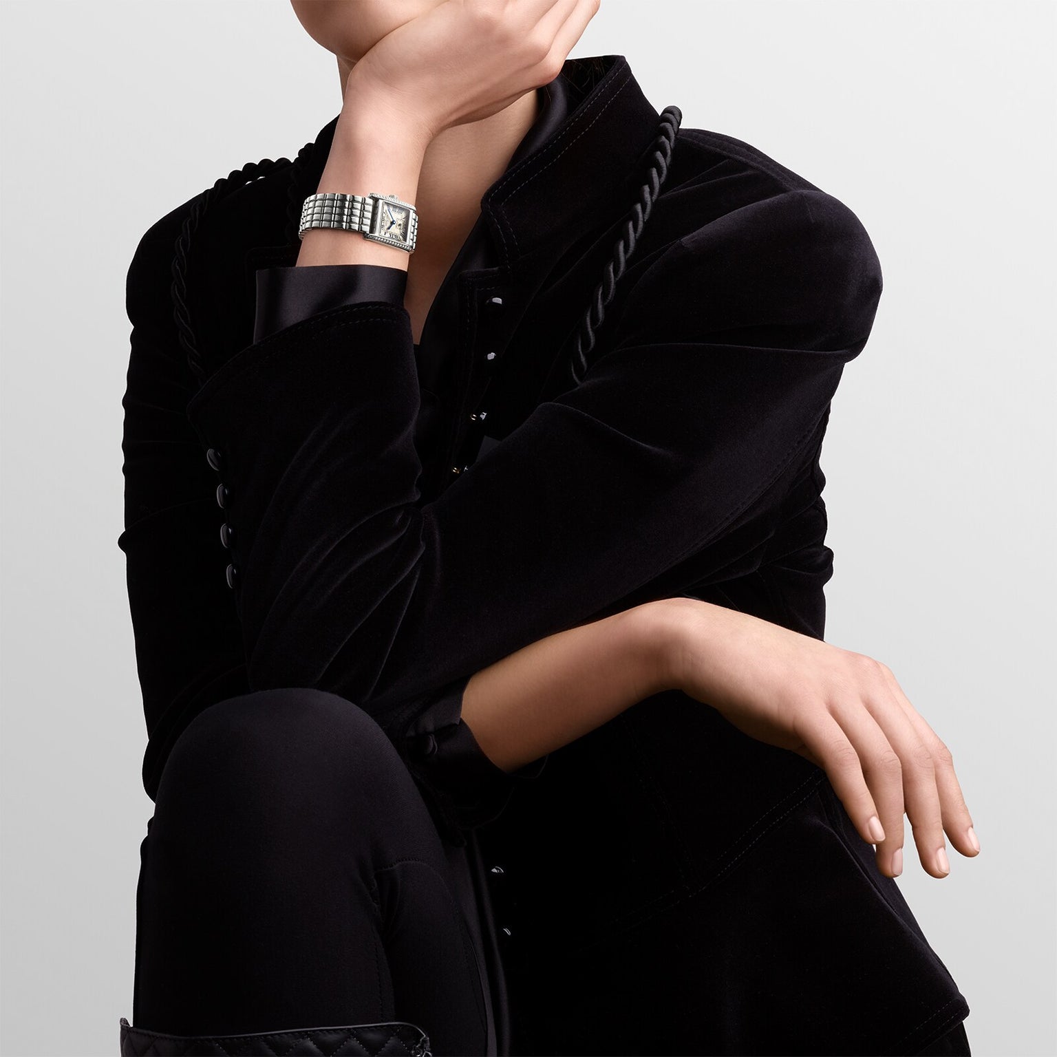 A person in a black velvet jacket and black pants wears a Longines Mini DolceVita watch with a stainless steel case, sitting with one hand on their knee and the other touching their chin.