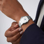 A person wearing the Longines Elegant Collection Moonphase, featuring a silver metal band, diamond indexes, and a white dial, adjusts it on their left wrist. The watch complements their dark long-sleeve garment.