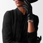 A stylish woman in black, wearing leather gloves, a wide-brimmed hat partially covering her face, completes her look with the Longines Dolcevite watch.