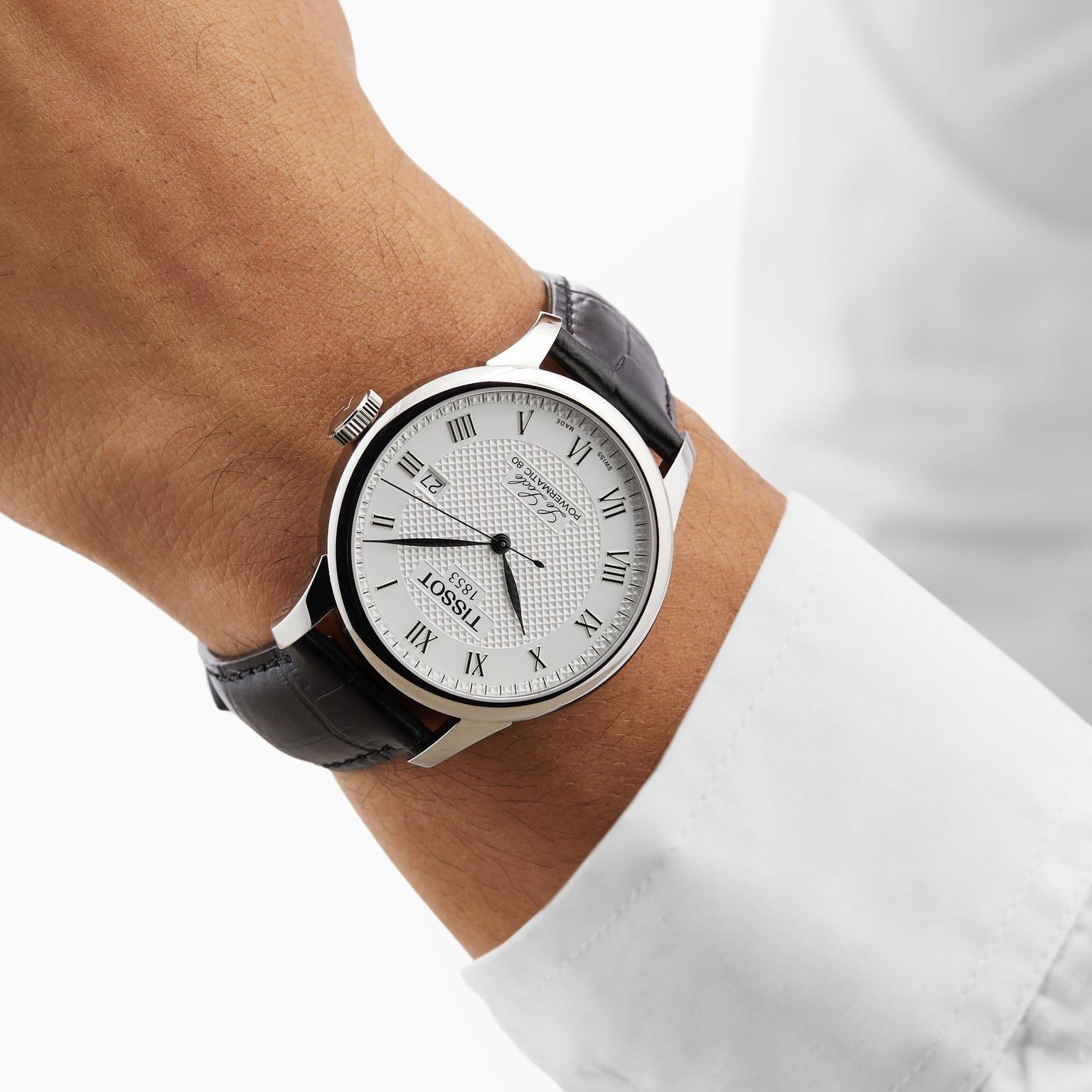 A person wearing a Tissot Le Locle Powermatic 80 with a white dial, black Roman numerals, and black leather strap, paired with a white long-sleeve shirt—a nod to classic Swiss style.