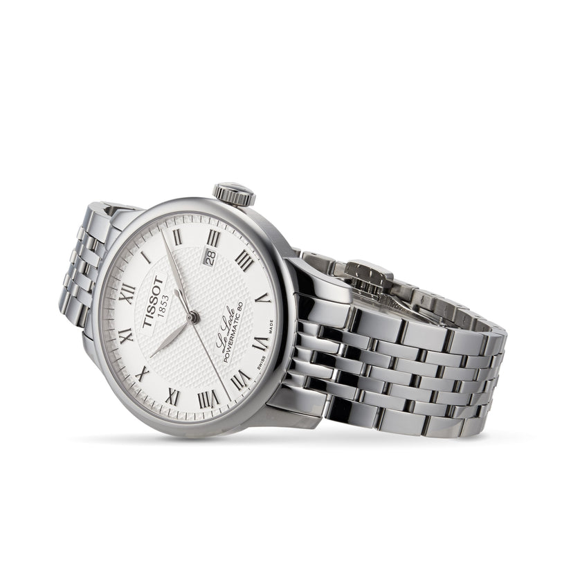 The Tissot Le Locle Powermatic 80 silver wristwatch features a white dial with Roman numerals, date display, and metal link band, presented on a white background. Inspired by the Swiss Jura Mountains.