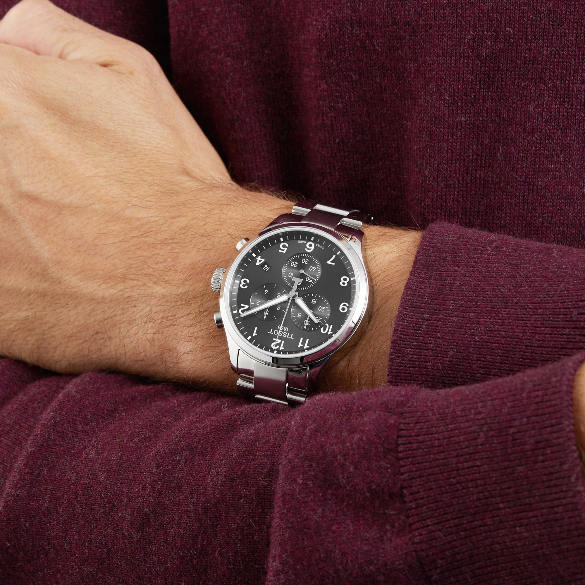 A person wearing a Tissot Chrono XL Classic wristwatch with a black face and Arabic numerals, visible over the sleeve of a burgundy sweater.