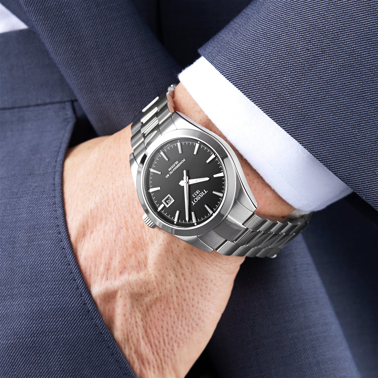 A person in a dark blue suit and white shirt wears the Tissot Gentleman Powermatic 80 Silicium watch, featuring a stainless steel case, black dial, and metal band.