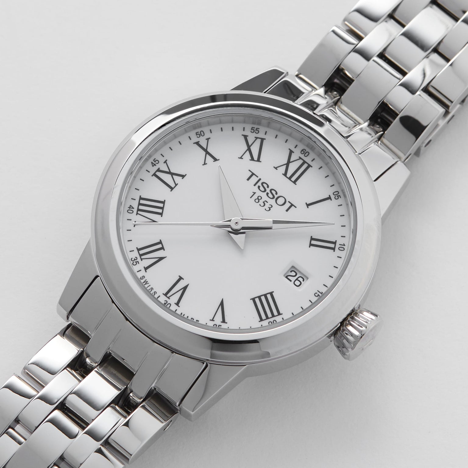 The Tissot Classic Dream features Roman numeral markers, a white dial, date window at 3 o’clock, and a metal link bracelet—Swiss quartz movement blends luxury and style, all showcased on a white background.