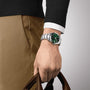 A person wearing a Tissot Gentleman Powermatic 80 Silicium watch with a green dial holds brown leather handles, dressed in tan pants and a black sweater with a white shirt cuff.