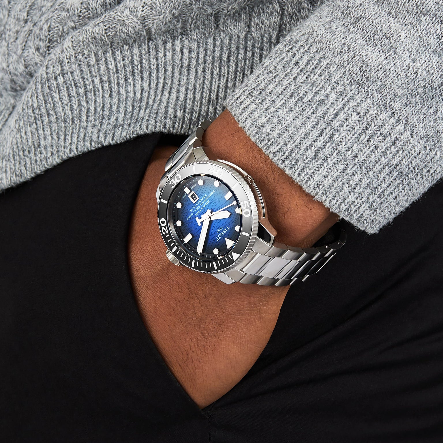 A person wearing a Tissot Seastar 2000 Professional Powermatic 80 watch with a blue face and black bezel, paired with a grey sweater and black pants.