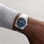 A person in a light gray long-sleeve shirt wears the Tissot PRX Powermatic 80—a luxurious silver wristwatch featuring a blue face, stainless steel case, and date display.