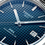 Close-up of the Tissot PRX Powermatic 80 watch, featuring a blue textured dial, silver hands and markers, stainless steel case, and a date window displaying 14.