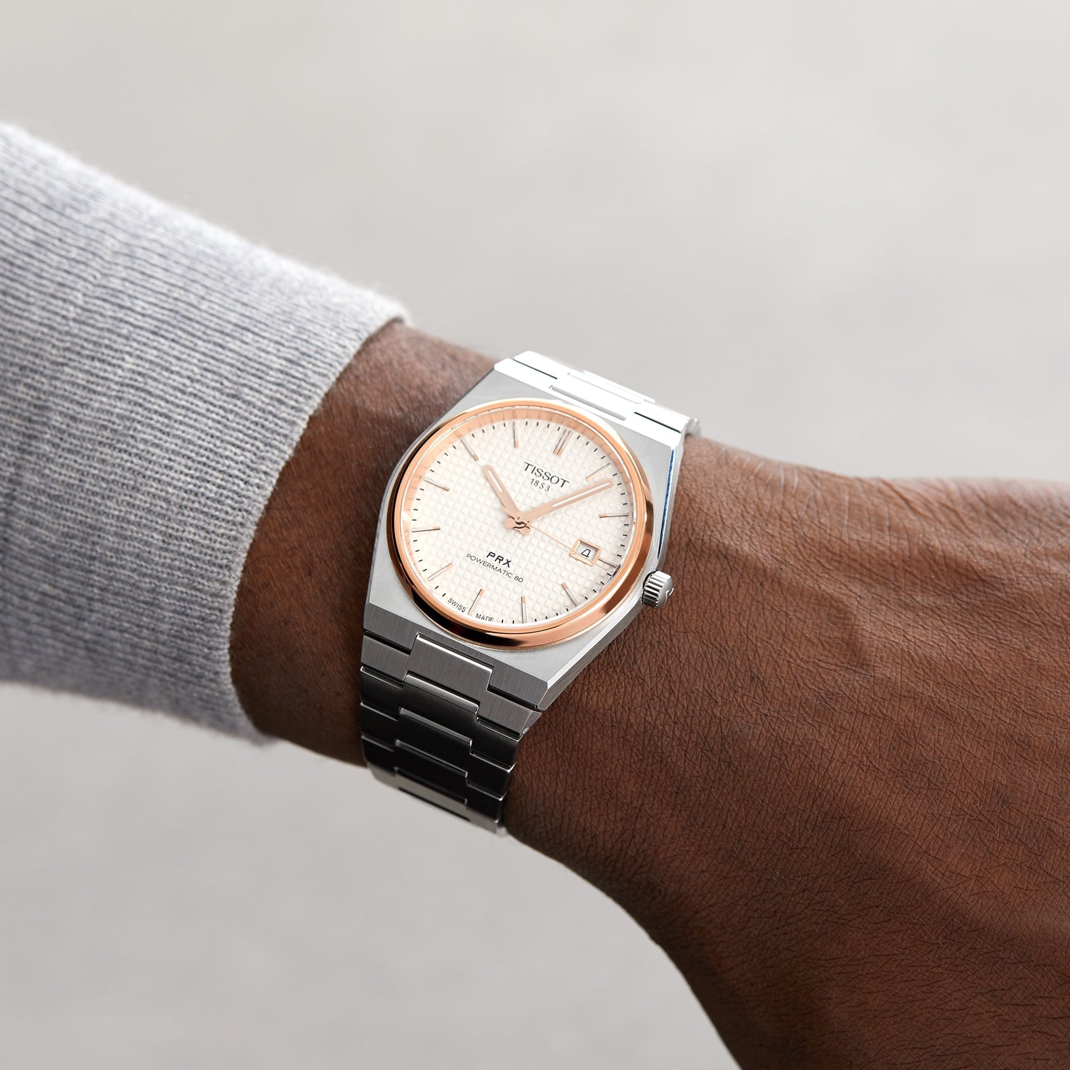 A person in a light grey long-sleeve top wears a Tissot PRX Powermatic 80 wristwatch with a silver stainless steel case, white dial, and date display.