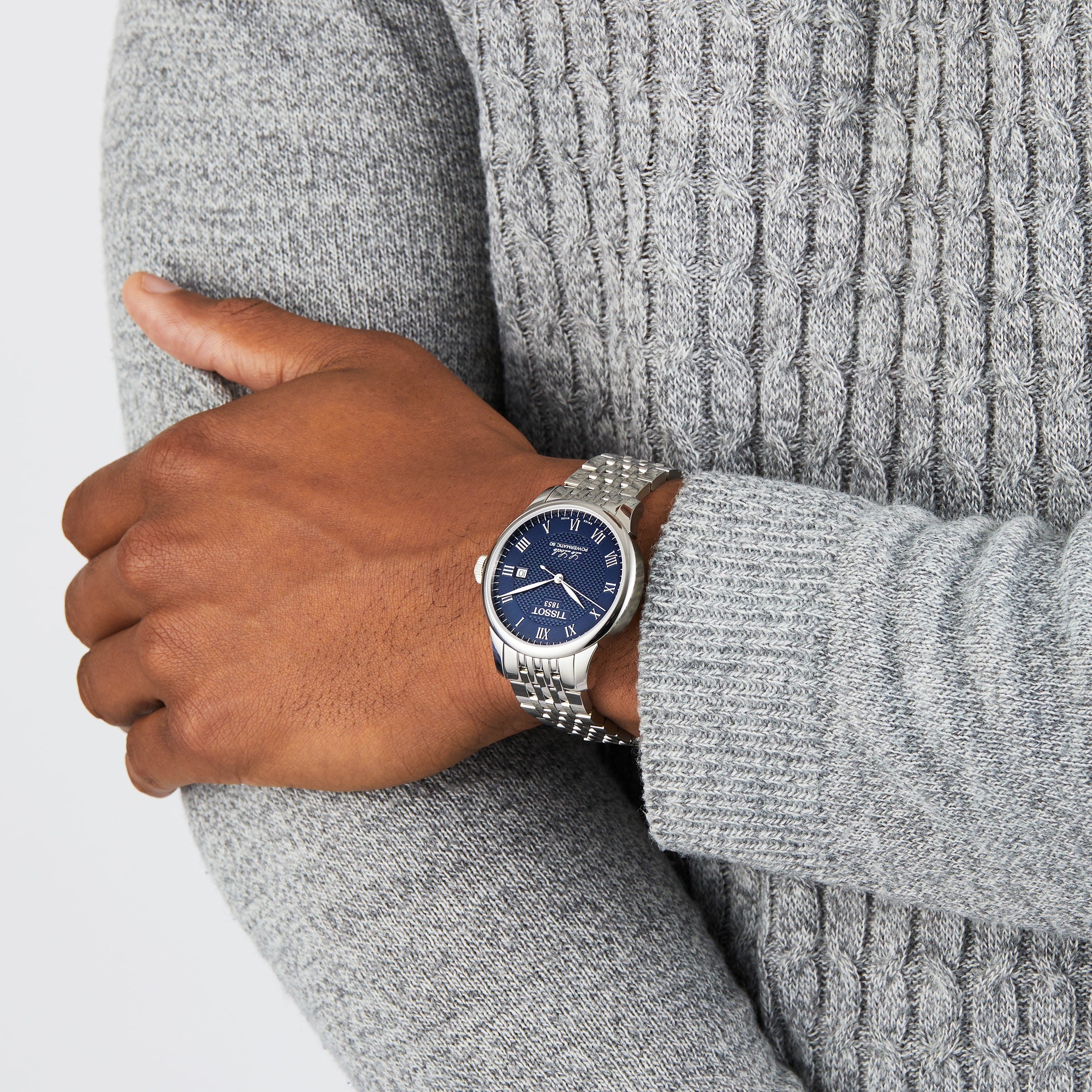 A person in a textured gray sweater displays a wrist with the Tissot Le Locle Powermatic 80, a silver watch featuring a blue dial, Roman numerals, and renowned Swiss precision.