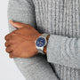 A person in a textured gray sweater displays a wrist with the Tissot Le Locle Powermatic 80, a silver watch featuring a blue dial, Roman numerals, and renowned Swiss precision.