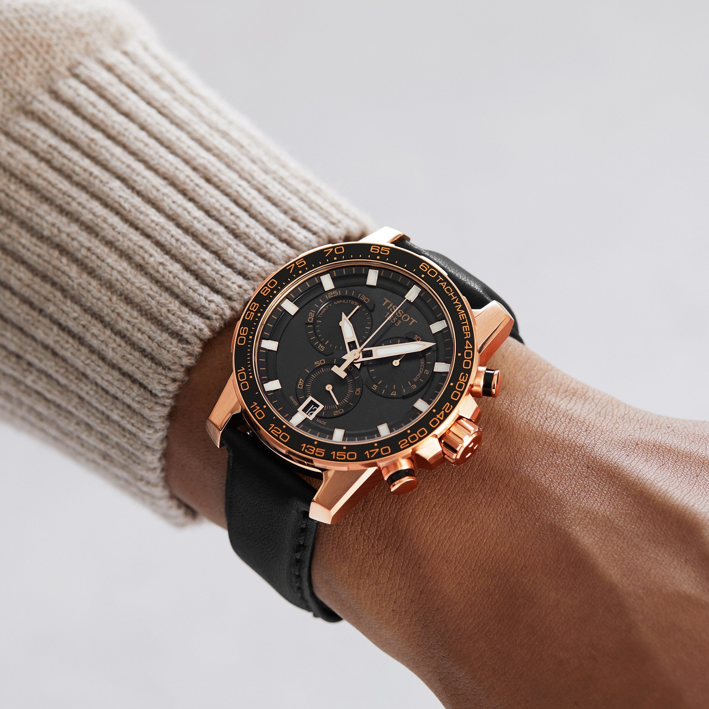 A person wearing a Tissot Supersport Chrono, featuring a rose gold case, black strap, and black dial.