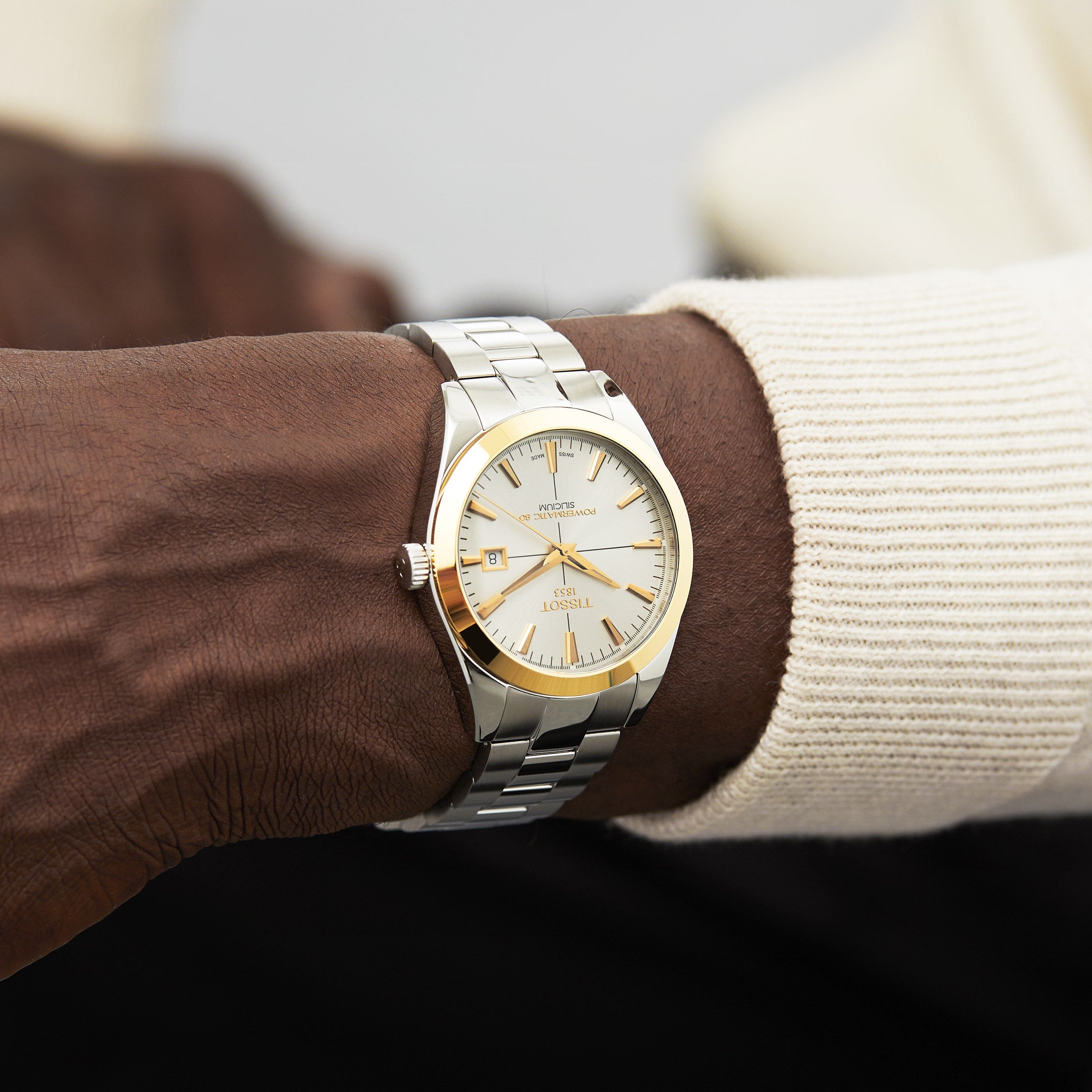 A close-up of a person wearing the Tissot Gentleman Powermatic 80 Silicium with a solid 18K gold bezel and white dial, paired with a cream-colored long sleeve.