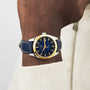 A person sports the Tissot Gentleman Powermatic 80 Silicium Solid 18k Bezel, featuring a blue face, gold accents, and a blue leather strap over a cream sweater—an effortless statement of luxury and style.