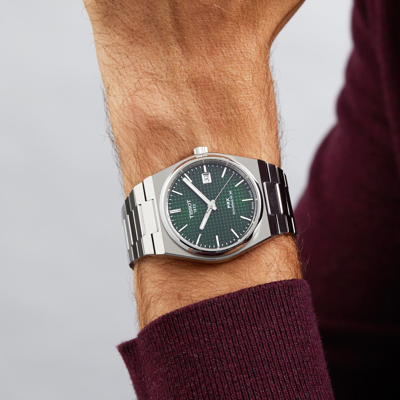 A Tissot PRX Powermatic 80 watch with a green dial and stainless steel case is worn on the left wrist, paired with a burgundy long-sleeve shirt.