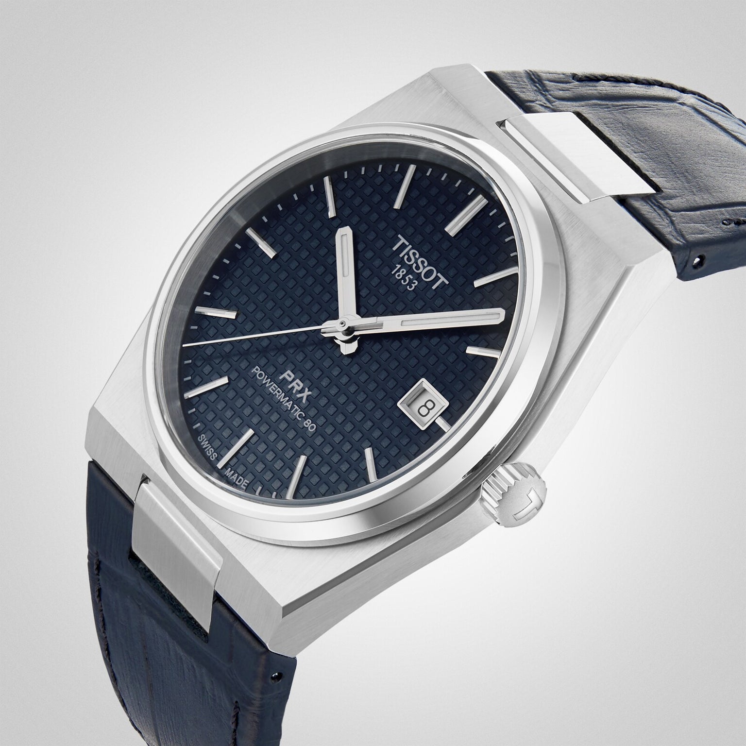 The Tissot PRX Powermatic 80 wristwatch features a blue textured dial, silver hour markers, date display, and a blue leather strap with a stainless steel case. Shown against a light background for an elegant appeal.