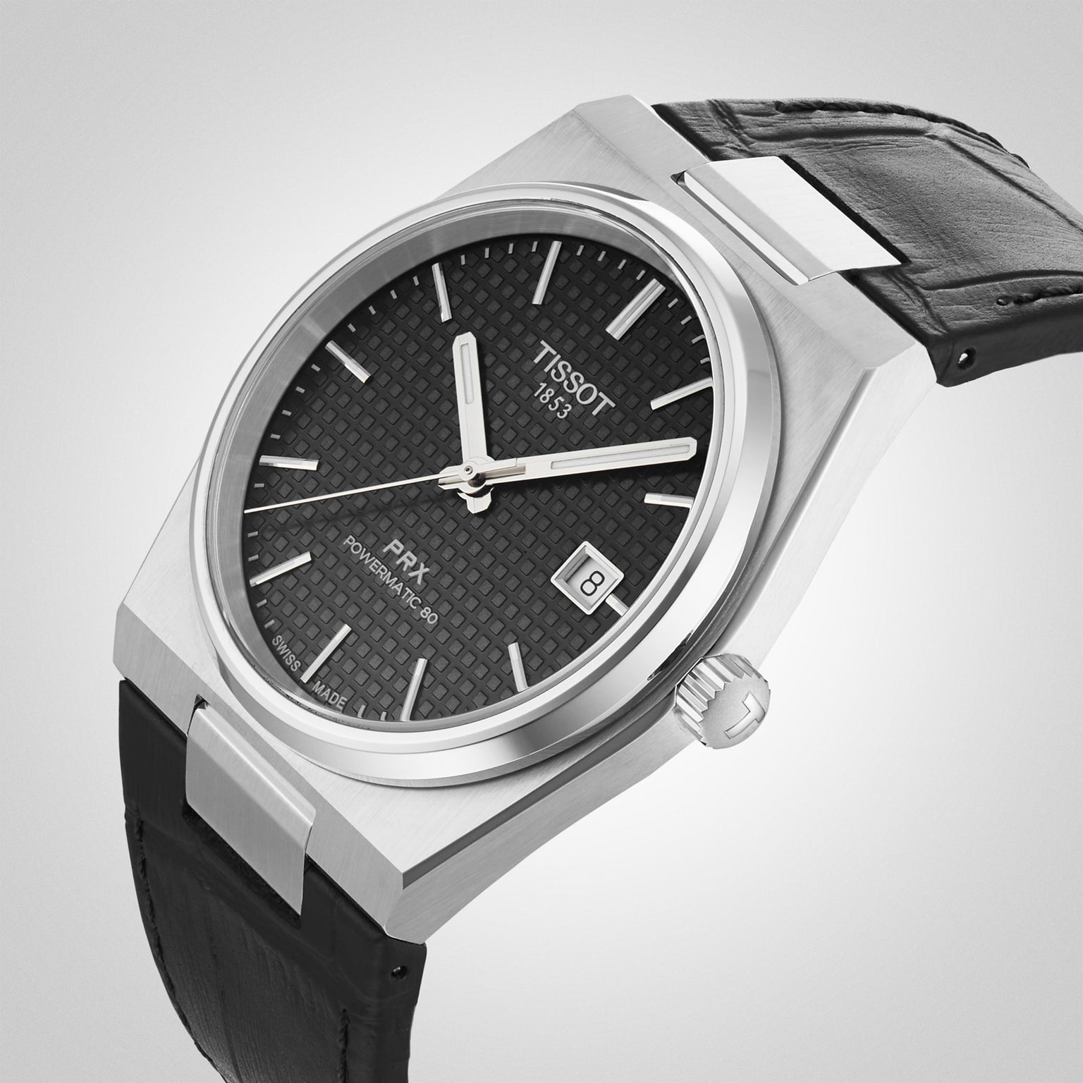 The Tissot PRX Powermatic 80 features a black textured dial, date display, silver hands, a black leather strap, and a stainless steel case with a 1970s-inspired design.