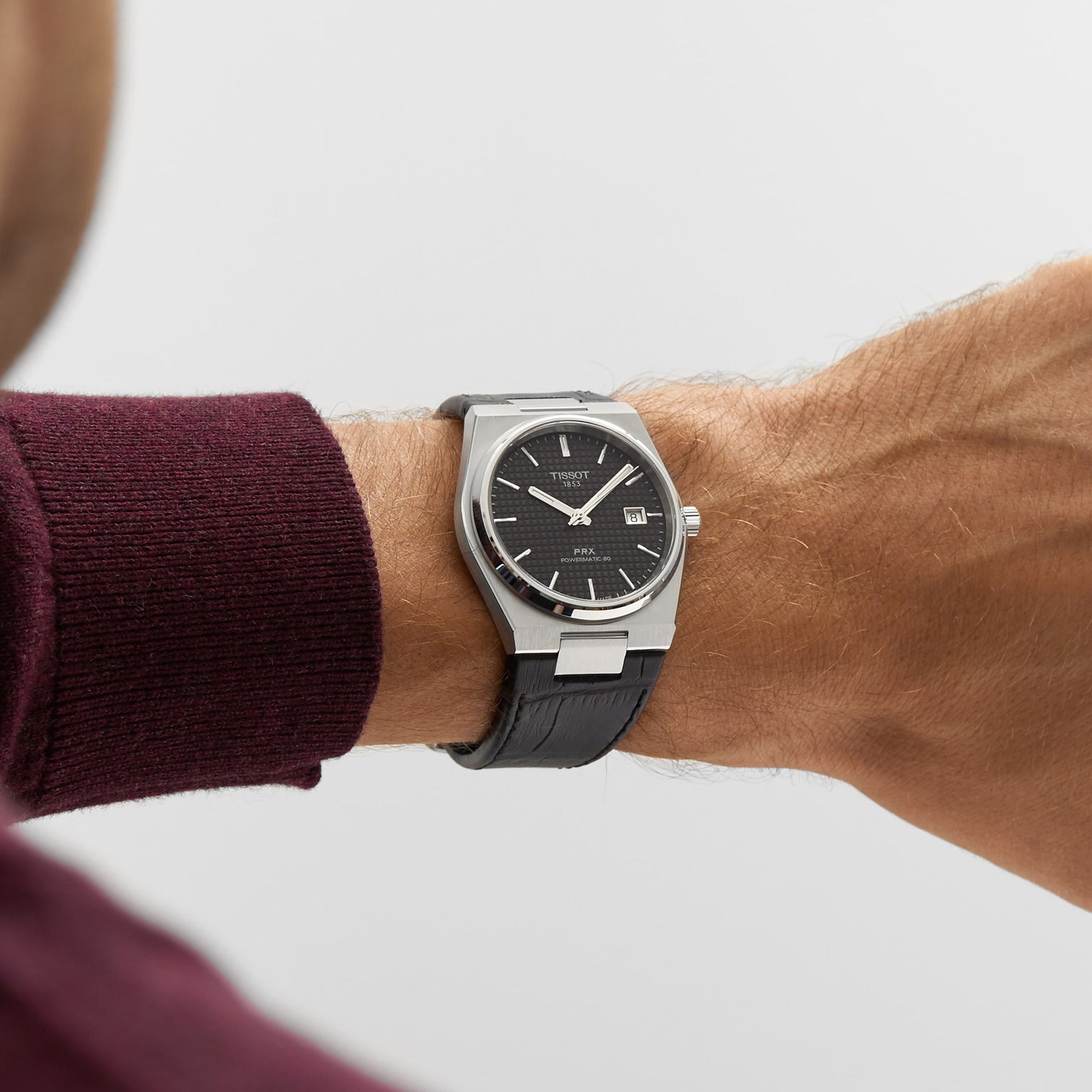 A person in a burgundy long-sleeve shirt wears the Tissot PRX Powermatic 80, featuring a stainless steel case, black dial, and black leather band.