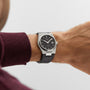 A person in a burgundy long-sleeve shirt wears the Tissot PRX Powermatic 80, featuring a stainless steel case, black dial, and black leather band.