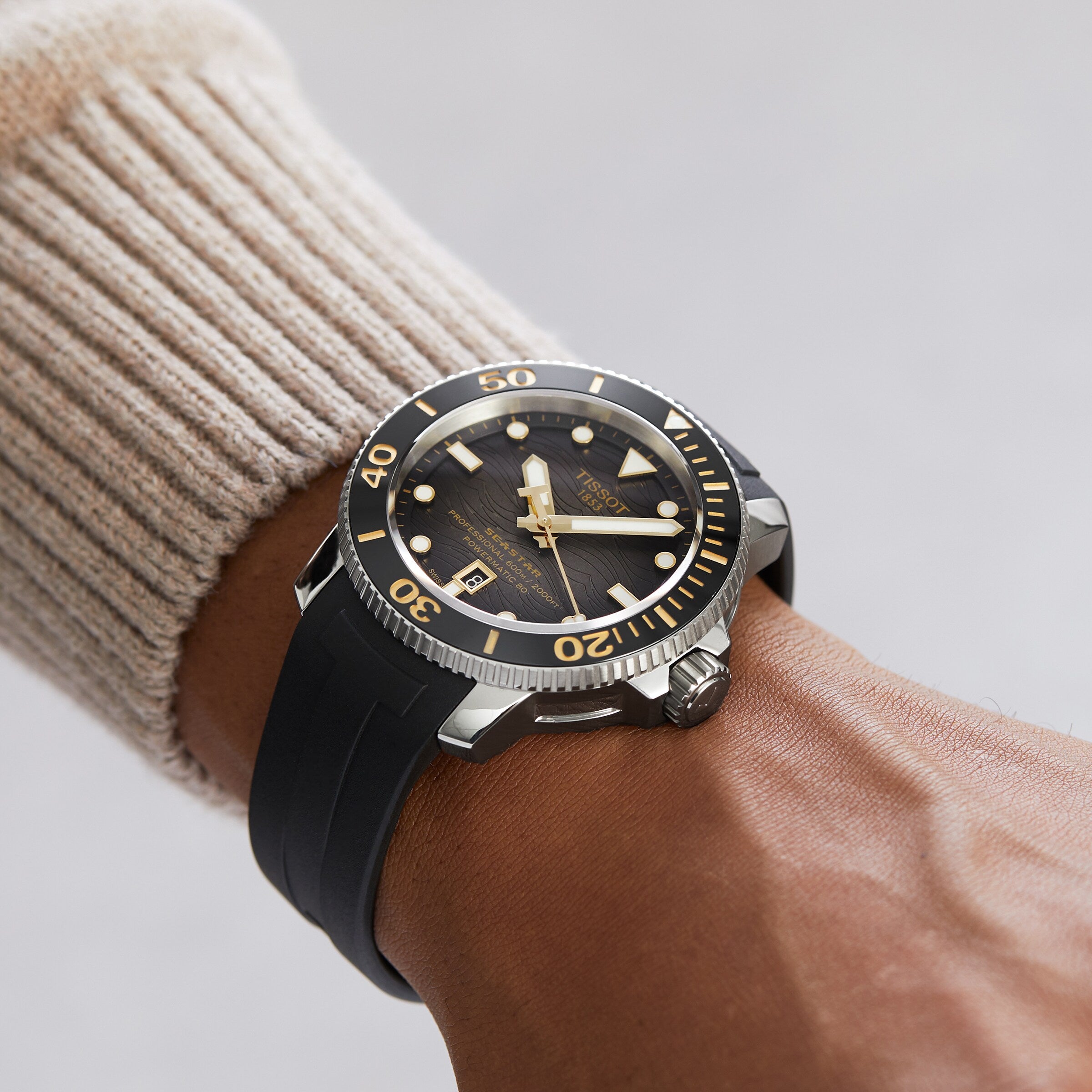 A person wears a Tissot Seastar 2000 Professional Powermatic 80—a black and gold diver’s watch with rotating bezel, date window, and black rubber strap—on their wrist.