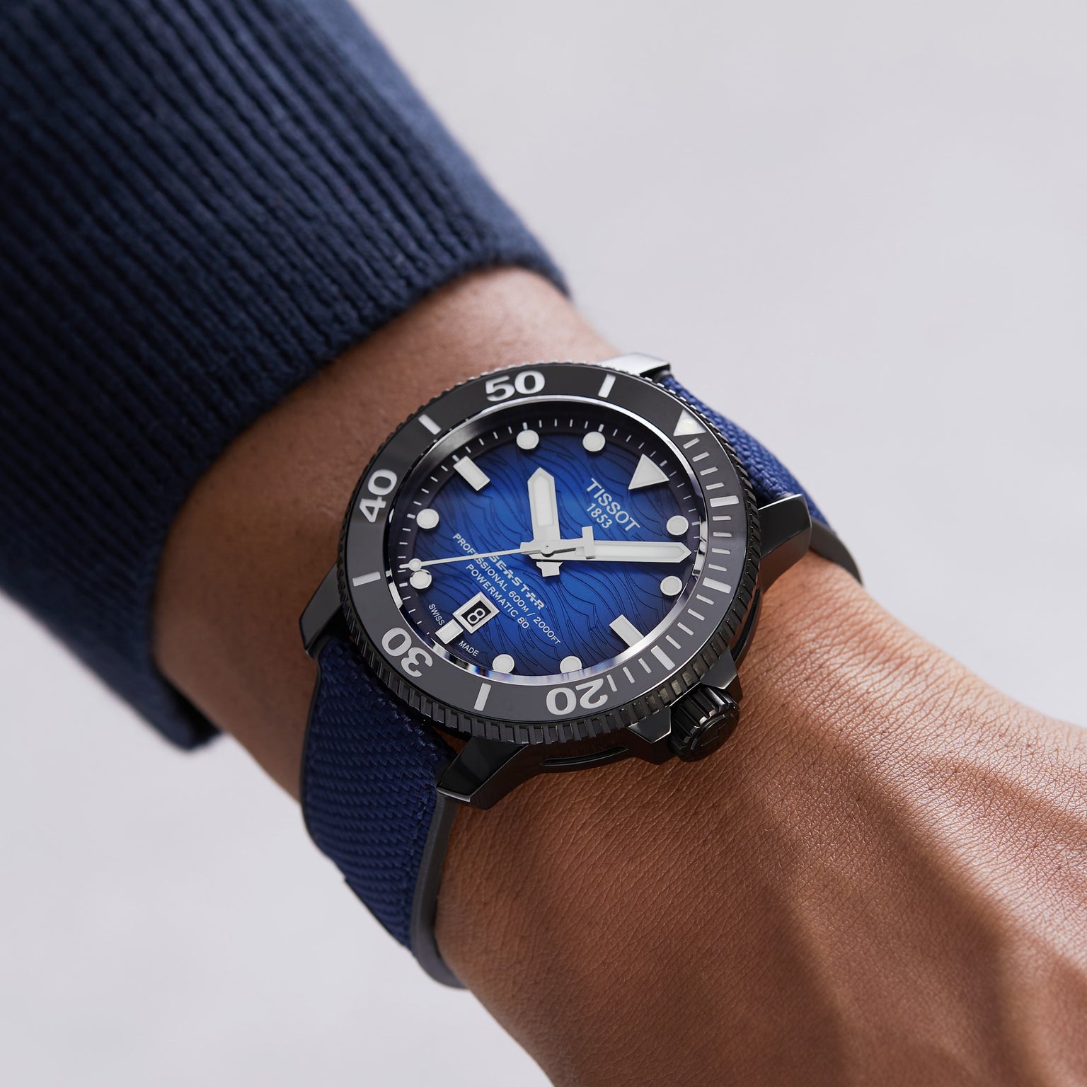 A person wearing a Tissot Seastar 2000 Professional Powermatic 80 with a black bezel and blue strap on their wrist, dressed in a dark blue long-sleeve shirt.