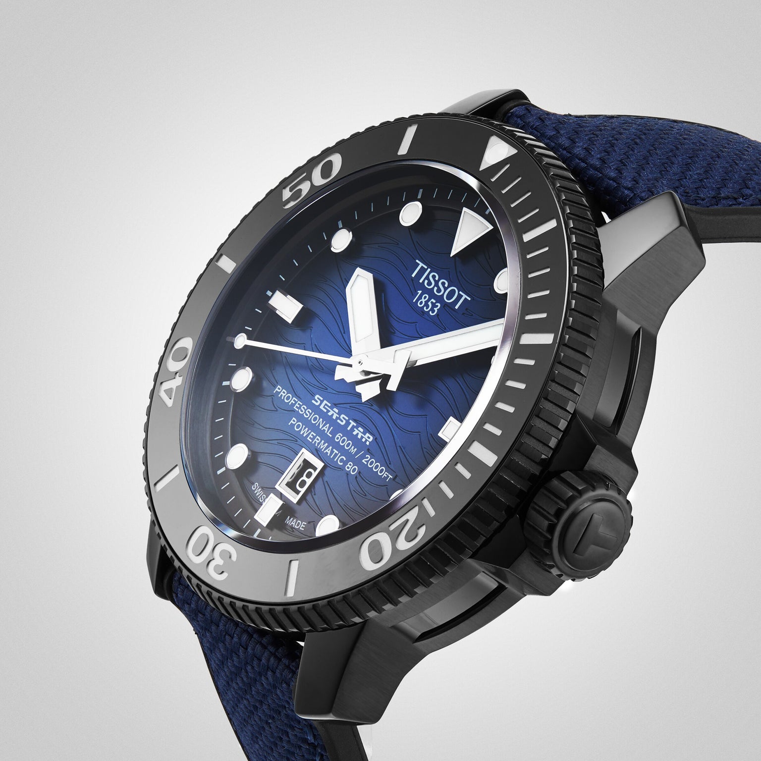 A close-up of the Tissot Seastar 2000 Professional Powermatic 80 shows its blue textured dial, black bezel, and blue fabric strap, displaying the time and date.