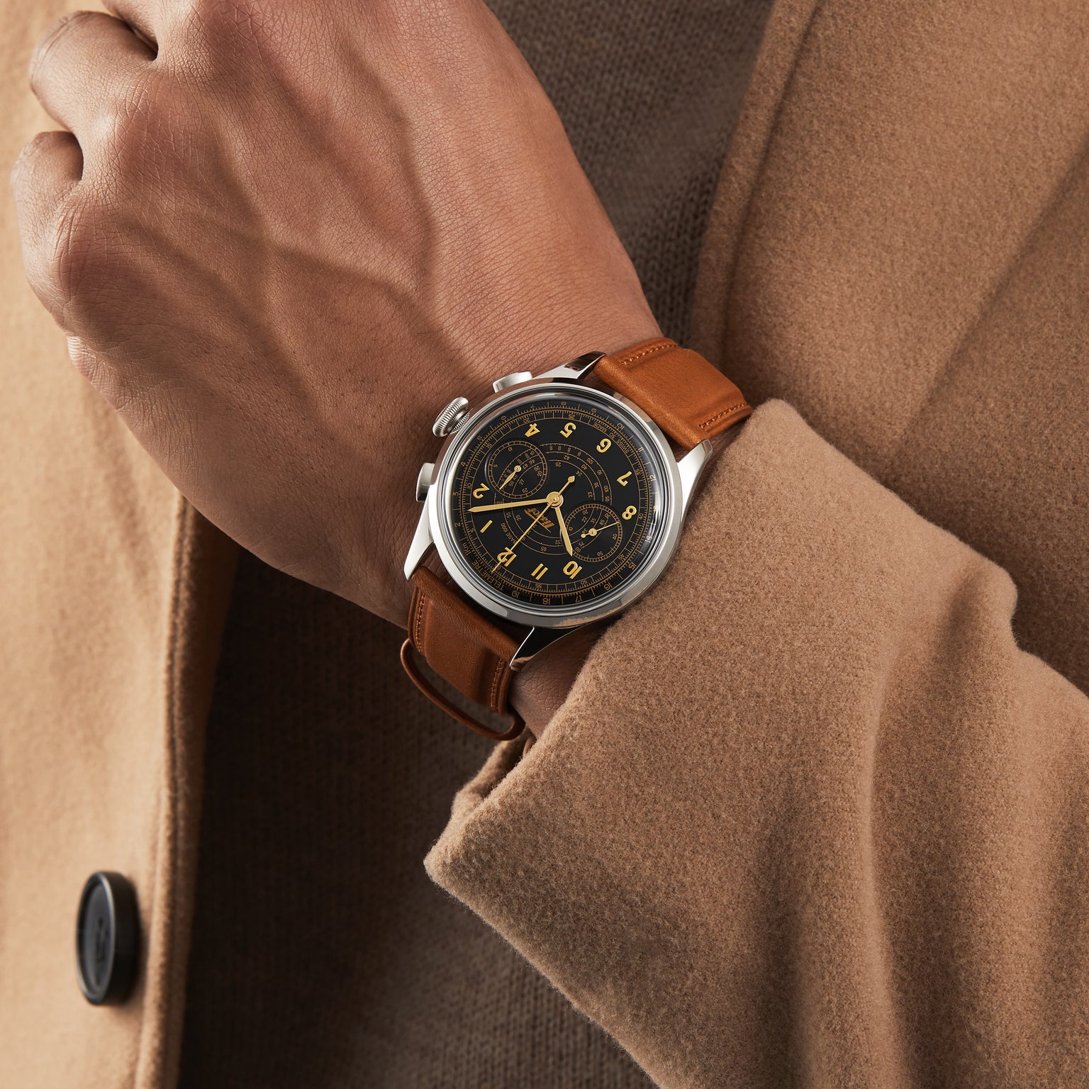 A person in a brown coat showcases the Tissot Telemeter 1938, which features a brown leather strap, black dial with gold numerals and chronograph subdials, and is powered by Swiss automatic movement.