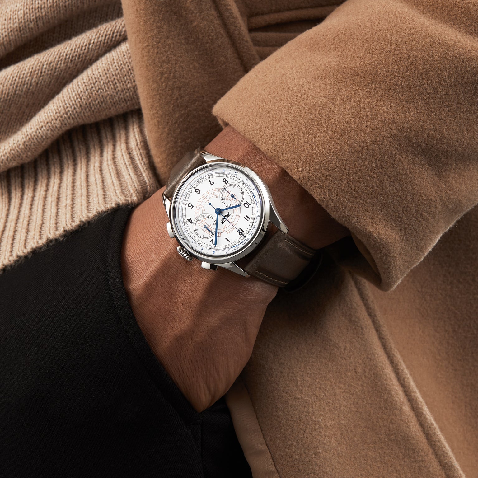 A person in a tan coat and black pants slips a hand into their pocket, showcasing the Tissot Telemeter 1938—a silver chronograph wristwatch with Swiss automatic movement and a brown leather strap that exudes refined vintage style.
