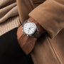 A person in a tan coat and black pants slips a hand into their pocket, showcasing the Tissot Telemeter 1938—a silver chronograph wristwatch with Swiss automatic movement and a brown leather strap that exudes refined vintage style.