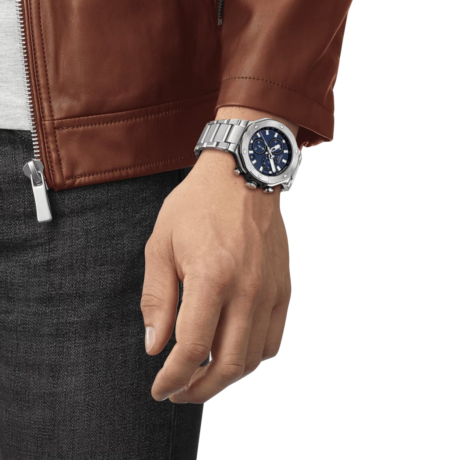 A person in a brown leather jacket and dark jeans wears a Tissot T-Race Chronograph, a motorcycle-inspired Swiss quartz watch with a blue face, on their left wrist.