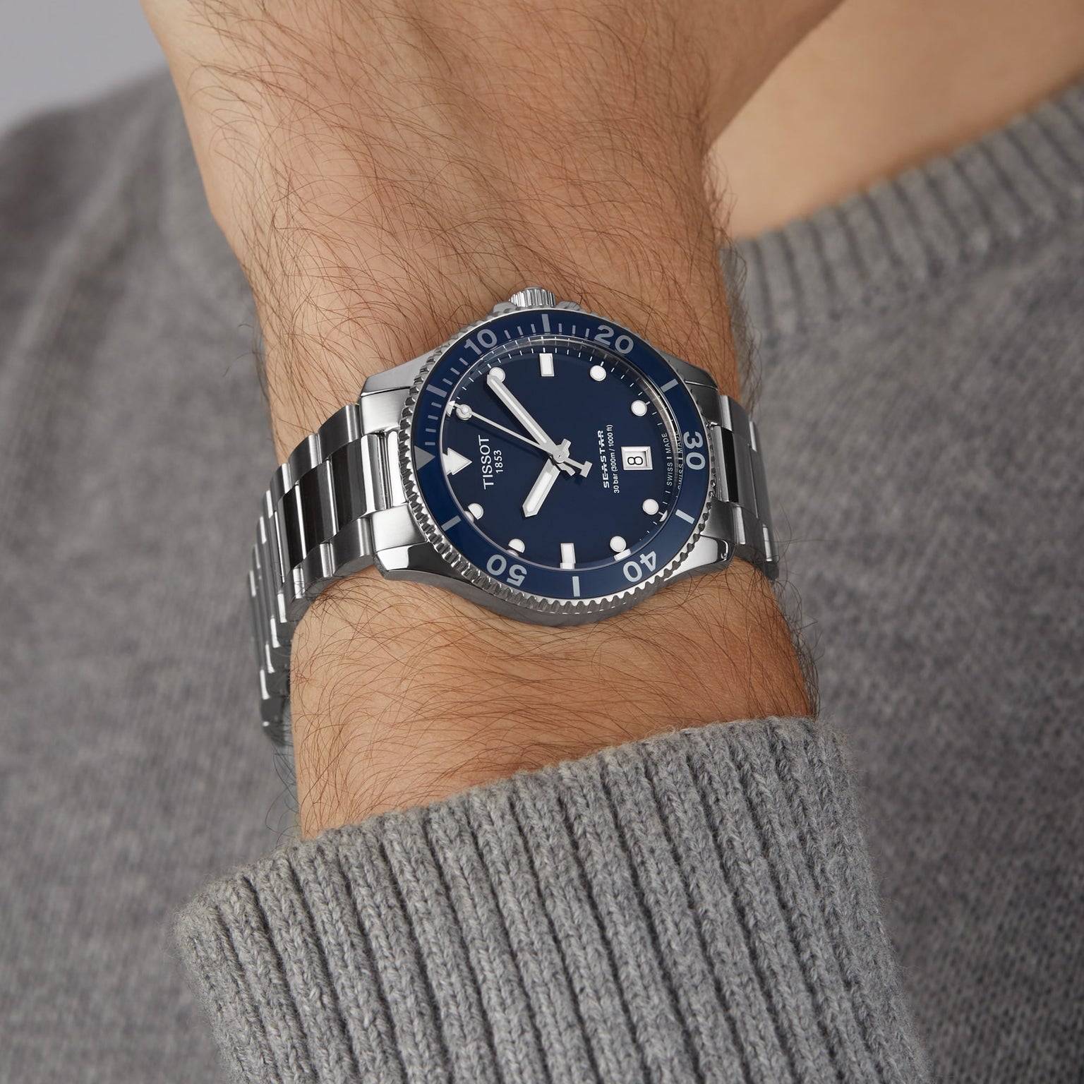 A person wears a Tissot Seastar 1000 silver metal diver’s watch with a blue face, bezel, and date display on their left wrist, paired with a gray knit sweater for a stylish look.