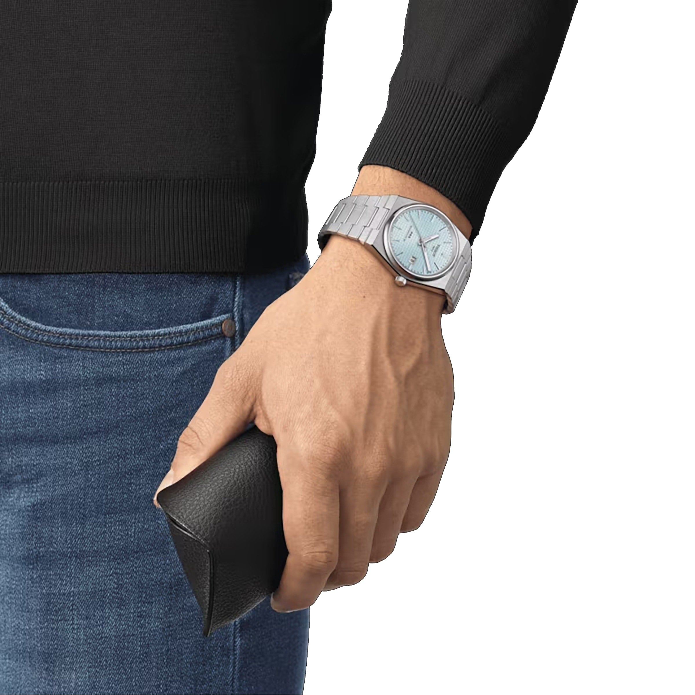 A person wearing a Tissot PRX Powermatic 80 with a stainless steel case, holding a black leather case, dressed in a black long-sleeve shirt and blue jeans.