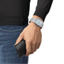A person wearing a Tissot PRX Powermatic 80 with a stainless steel case, holding a black leather case, dressed in a black long-sleeve shirt and blue jeans.